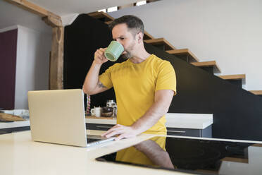 Mann trinkt Kaffee und benutzt einen Laptop auf einer Kücheninsel zu Hause - MTBF00470