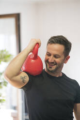 Nahaufnahme eines lächelnden reifen Mannes, der zu Hause eine Kettlebell auf der Schulter hält - MTBF00482