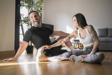 Cheerful man sitting with woman exercising on hardwood floor at home - MTBF00485