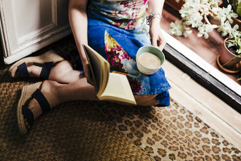 Woman holding coffee cup and book while sitting on floor at home - EBBF00300