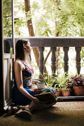 Woman holding cup and book while sitting by balcony at home - EBBF00301