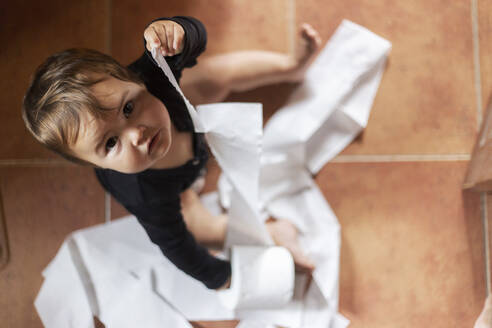 Von oben aufgenommenes Bild eines süßen Babys, das auf dem Boden sitzend Toilettenpapier abrollt und in die Kamera schaut - ADSF00045