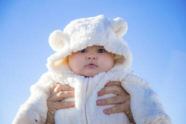 Hände einer anonymen Person, die ein bezauberndes Baby in warmer Kleidung vor einem wolkenlosen blauen Himmel hält - ADSF00051