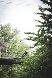 Mature man with sword sitting on old pipes against sky - HMEF00962