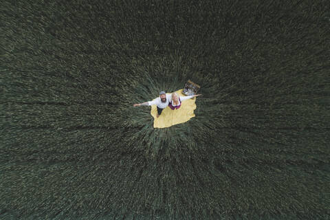 Drone shot of mature couple with arms outstretched standing on blanket amidst green cornfield stock photo