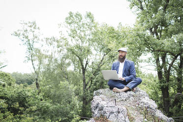 Geschäftsmann in Anzug und Mütze, der einen Laptop benutzt, während er auf einem Felsen vor Bäumen sitzt - HMEF00986