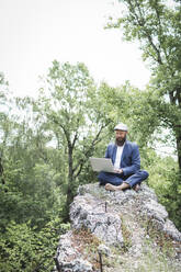Geschäftsmann im Anzug arbeitet an einem Laptop, während er auf einem Felsen vor Bäumen sitzt - HMEF00987