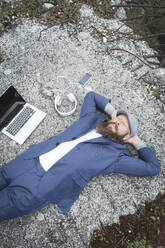 Bearded businessman with hands behind head relaxing on land in forest - HMEF00992