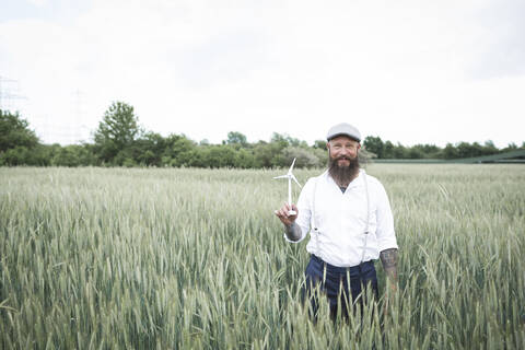 Bärtiger Mann, der eine kleine Windmühle hält, während er inmitten eines Kornfelds vor dem Himmel steht, lizenzfreies Stockfoto