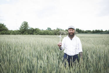 Bearded man holding small windmill while standing amidst cornfield against sky - HMEF01006