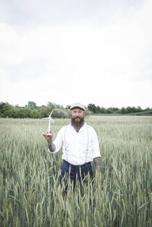 Mature man holding small windmill while standing amidst cornfield against sky - HMEF01007