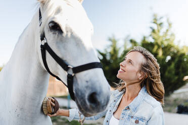 Smiling woman brushing a horse on a farm - MRRF00091