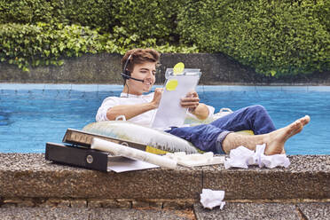 Junger Mann sitzt auf einer Luftmatratze im Schwimmbad und arbeitet - UKOF00001