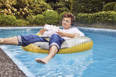 Young man working from home office on airbed in swimming pool looking at architectural model - UKOF00003