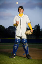 Porträt eines Jungen in weißer Baseballuniform und Schienbeinschützern, der einen Baseball in die Luft wirft - CAVF86228