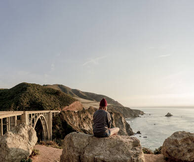 Frau sitzt auf einem Felsen mit Blick auf die Küste von Big Sur und Bixby Br - CAVF86308