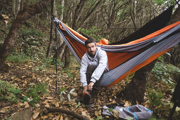 Ein Mann sitzt in einer Hängematte im Wald und bereitet sich auf eine Wanderung vor - CAVF86341