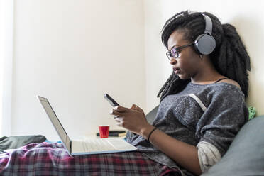 Junge Frau, die ein Smartphone benutzt, mit Laptop und Kopfhörern auf dem Bett sitzend, zu Hause - MEUF01048