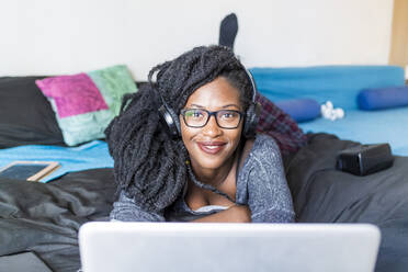 Young woman using laptop and headphones on bed at home - MEUF01051