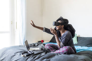 Junge Frau mit Virtual-Reality-Brille auf dem Bett sitzend - MEUF01055