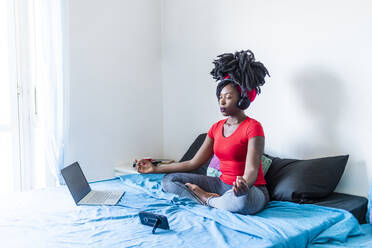 Junge Frau meditiert am Laptop auf dem Bett sitzend - MEUF01064