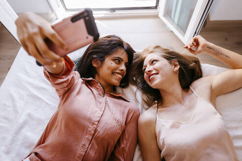 Friends looking at each other while taking selfie on bed at home - TCEF00806