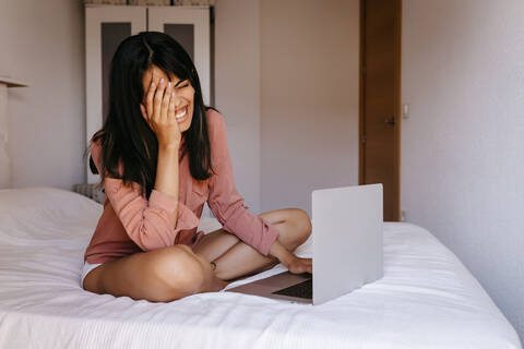 Junge Frau benutzt einen Laptop, während sie zu Hause auf dem Bett sitzt, lizenzfreies Stockfoto