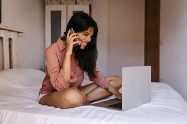 Lächelnde junge Frau, die zu Hause ein Mobiltelefon und einen Laptop benutzt - TCEF00809