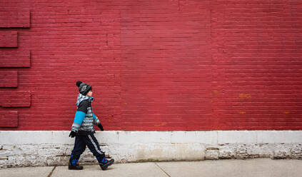 Young boy in winter clothing walking on sidewalk in front of red