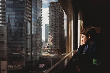Tween boy looking out a window at tall buildings of the city outside. - CAVF86405