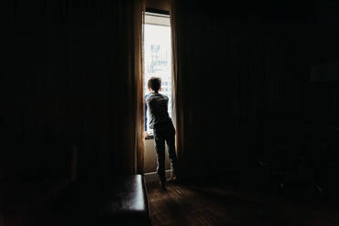 Young boy looking out a window of a darkened room at tall buildings. - CAVF86408