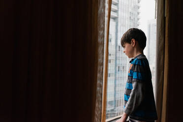 Young boy looking out a window at tall buildings of the city outside. - CAVF86410