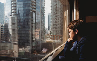 Tween boy looking out a window at tall buildings of the city outside. - CAVF86411