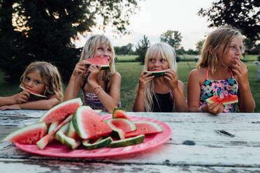Geschwister sitzen draußen am Picknicktisch und essen Wassermelone im Sommer - CAVF86495
