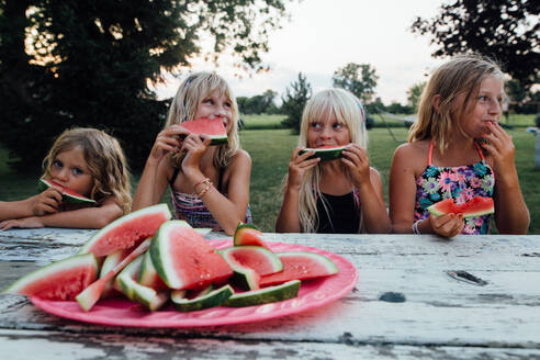 Geschwister sitzen draußen am Picknicktisch und essen Wassermelone im Sommer - CAVF86495