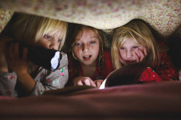 Drei kleine Mädchen lesen ein Buch unter einer Decke mit Taschenlampe - CAVF86503