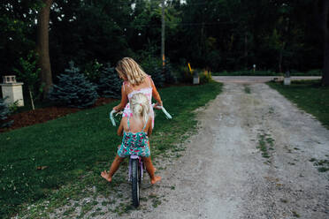 Two girls riding a bike down a driveway in the summer - CAVF86513