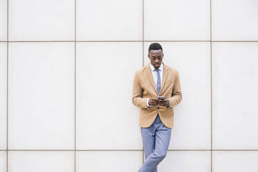 Young businessman leaning against a wall using smartphone - ABZF03216