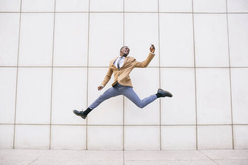 Young businessman jumping and taking a selfie in front of a wall - ABZF03218