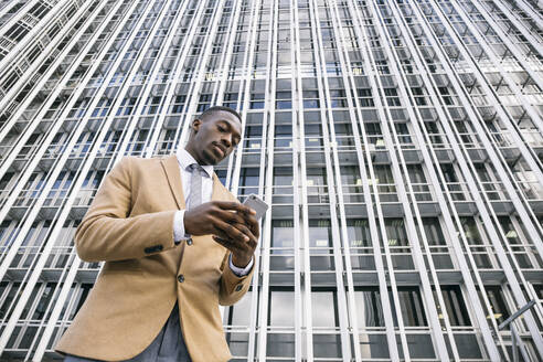 Young businessman in front of modern office building in the city using smartphone - ABZF03227