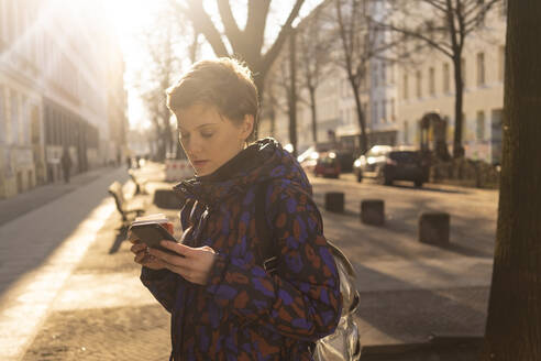Portrait of woman with backpack and coffee to go looking at cell phone in the evening, Berlin, Germany - TAMF02438