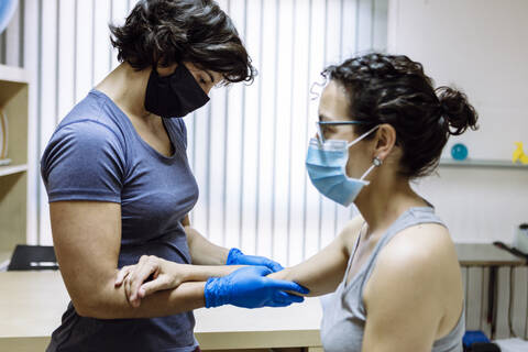 Physiotherapeutin mit Maske behandelt eine Kundin am Ellbogen, lizenzfreies Stockfoto