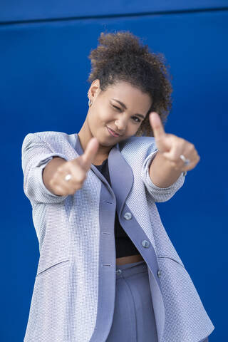 Smiling businesswoman with thumbs up in front of blue wall stock photo