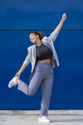 Smiling businesswoman with raised arm in front of blue wall - SNF00441