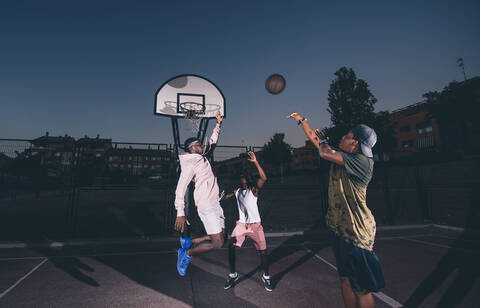 Männliche Freunde spielen Basketball im Gericht gegen klaren Himmel bei Nacht, lizenzfreies Stockfoto