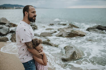 Tochter lehnt den Kopf an die Hand des Vaters, während sie am Strand steht - GMLF00320