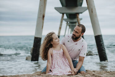 Father and daughter sitting on sand against pier at beach - GMLF00325