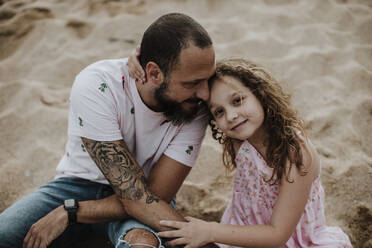 Lächelnde Tochter sitzt mit Vater am Strand im Sand - GMLF00327