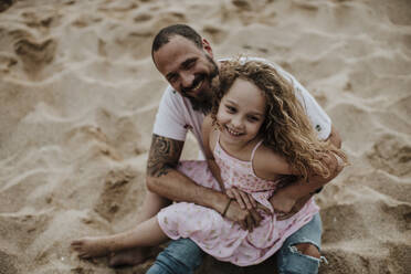 Daughter sitting on father's lap while playing at beach - GMLF00328