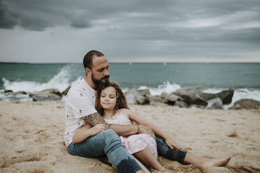 Father embracing daughter while sitting on beach - GMLF00332